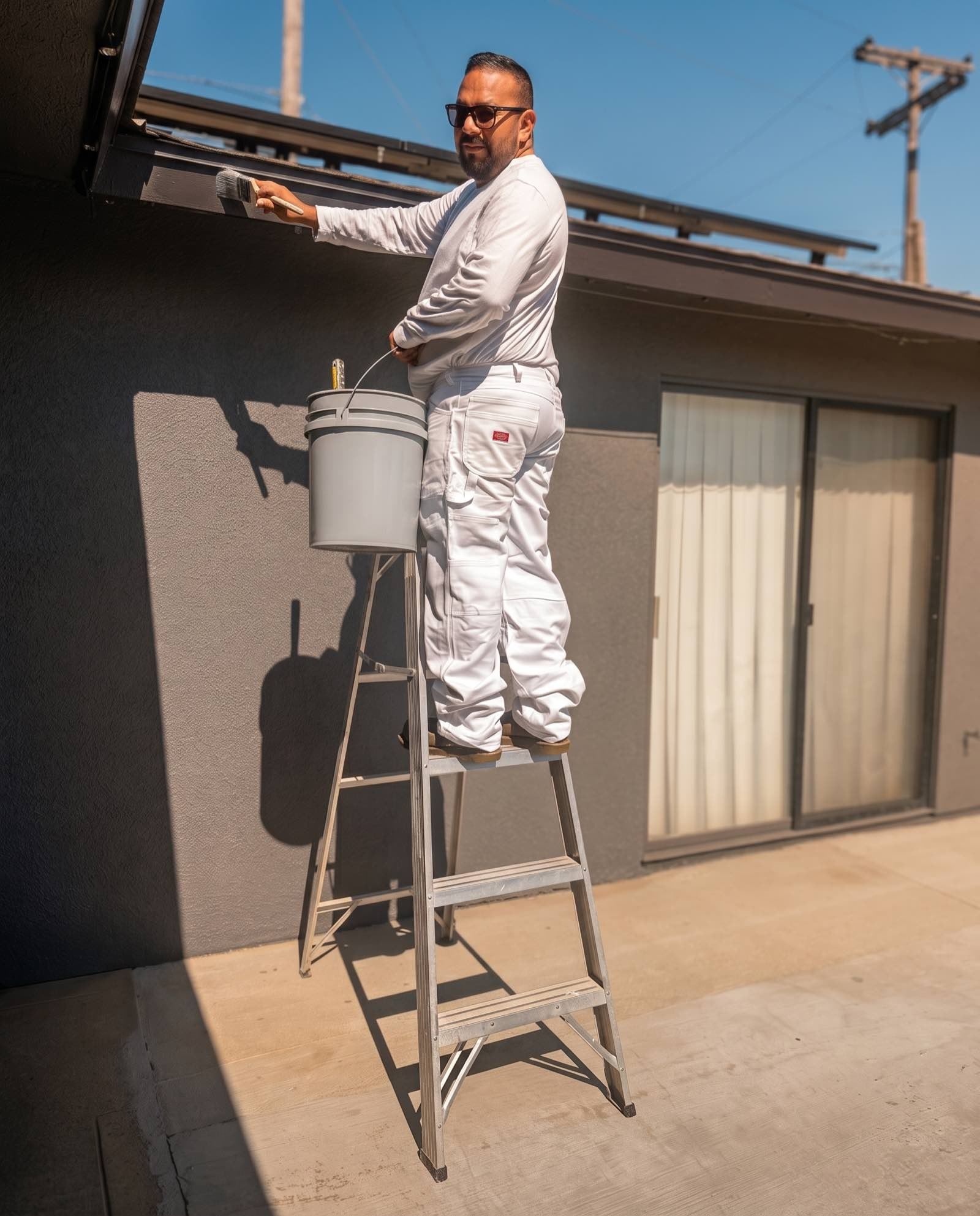 Pedro Ruiz, in painter's whites on a stepladder, painting the trim board along the roof edge of a single-storey house with a hand brush.