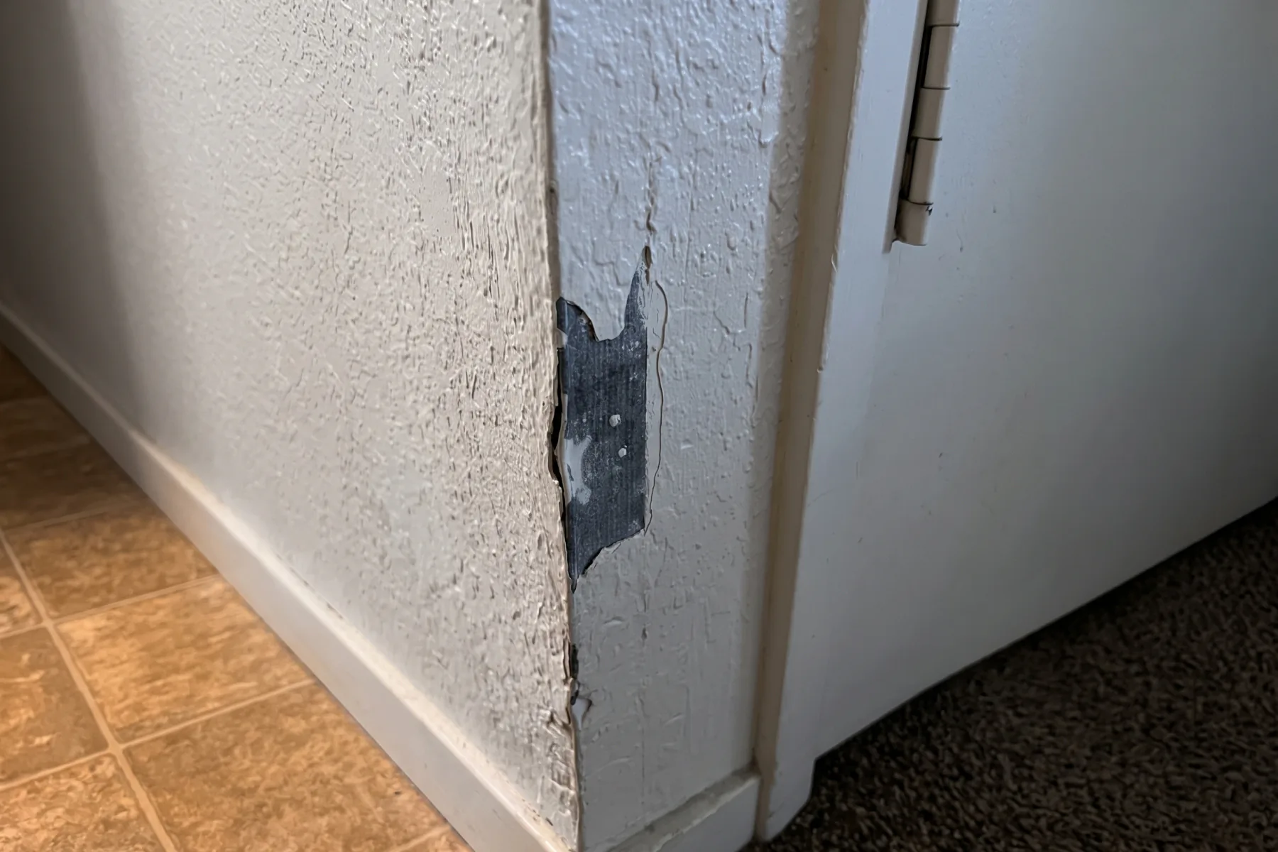 Before repair: an interior wall corner beside a doorway in Merced where a knock has taken off the paint and surface texture, exposing the dark board underneath, with a crack running up from the damage and the paint edges lifting.