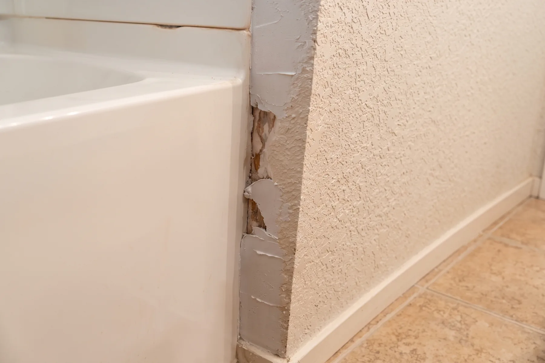 Before repair: the wall corner beside a bath in Merced with the surface broken away in a strip from top to bottom, exposing bare board and old patching compound, the torn edges lifting away from the wall.