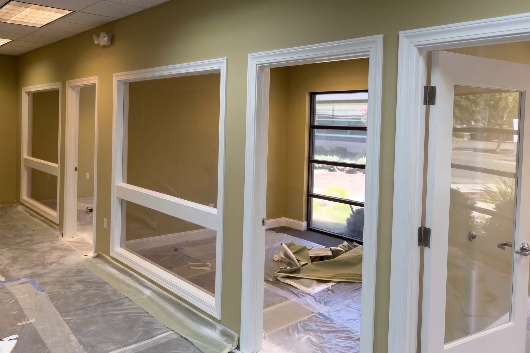 Before: an office corridor in Merced painted dark olive, with white window frames and door frames, the floor covered end to end in plastic sheeting taped down at the seams and a drop cloth through the open doorway.